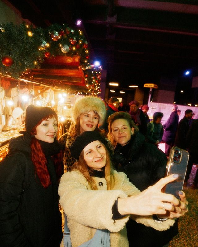 Fünf Frauen machen ein Selfie auf einem Weihnachtsmarkt, im Hintergrund leuchtende Lichter und Weihnachtsdekoration.
