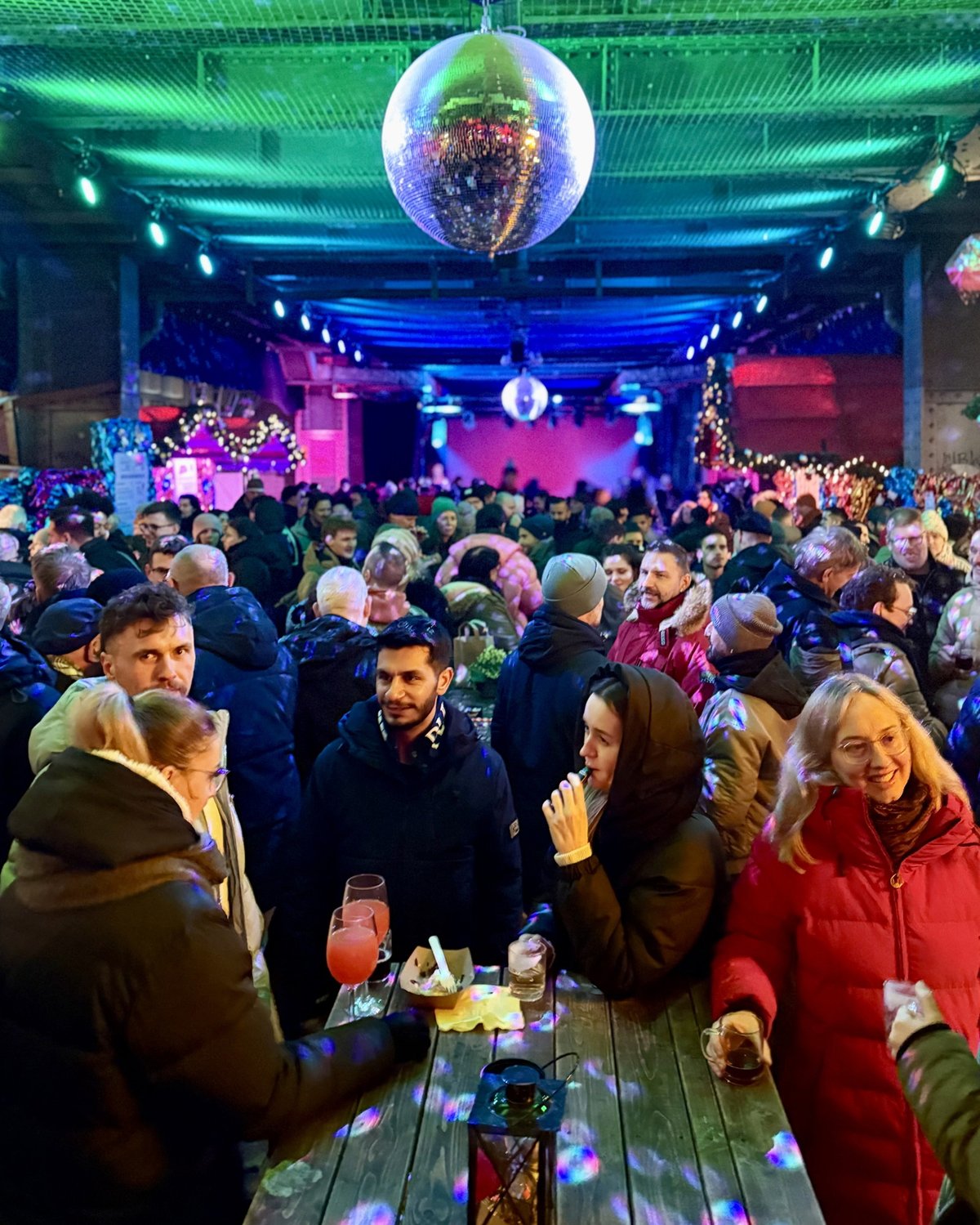Fröhliche Gäste stossen an einem Holztisch unter einer Discokugel an – Christmas Avenue Berlin
