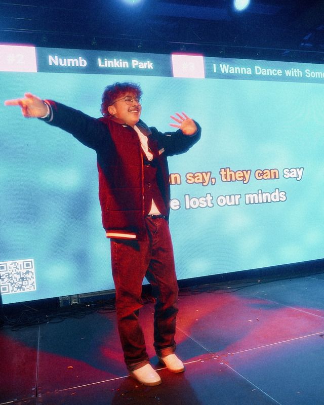 Ein junger Mann singt Karaoke vor einer großen Leinwand mit Text und Liedauswahl.