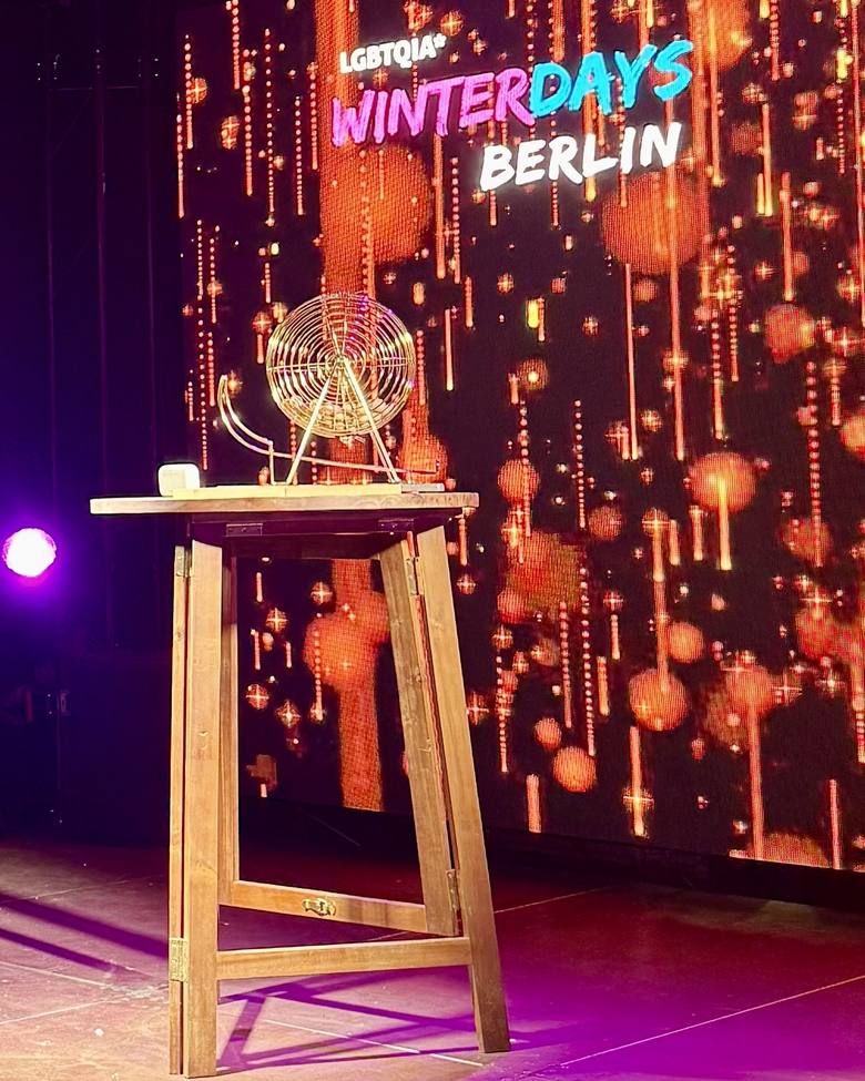 Eine goldene Bingotrommel steht auf einem hölzernen Stehtisch vor einem beleuchteten Bildschirm mit dem Schriftzug LGBTQIA* Winterdays Berlin.
