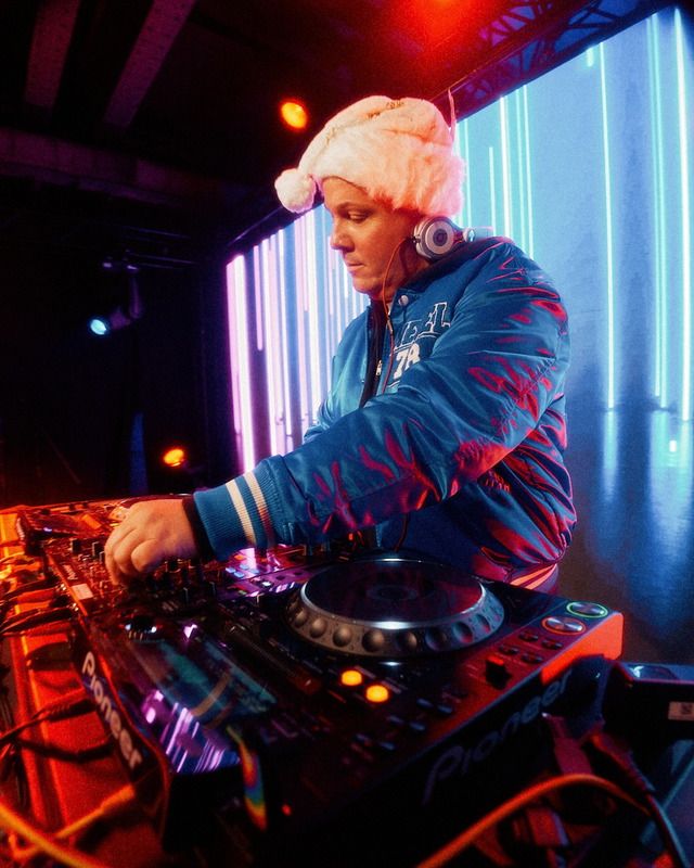 Ein DJ mit einer flauschigen Weihnachtsmütze steht an einem Mischpult und sorgt in einer beleuchteten Eventlocation für Stimmung.