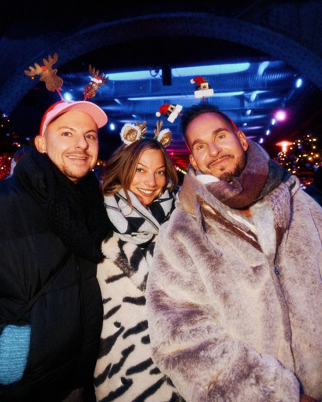 Drei fröhliche Gäste tragen weihnachtliche Haarreifen mit Rentiergeweihen und kleinen Weihnachtsmützen bei einer feierlichen Abendveranstaltung im Freien.