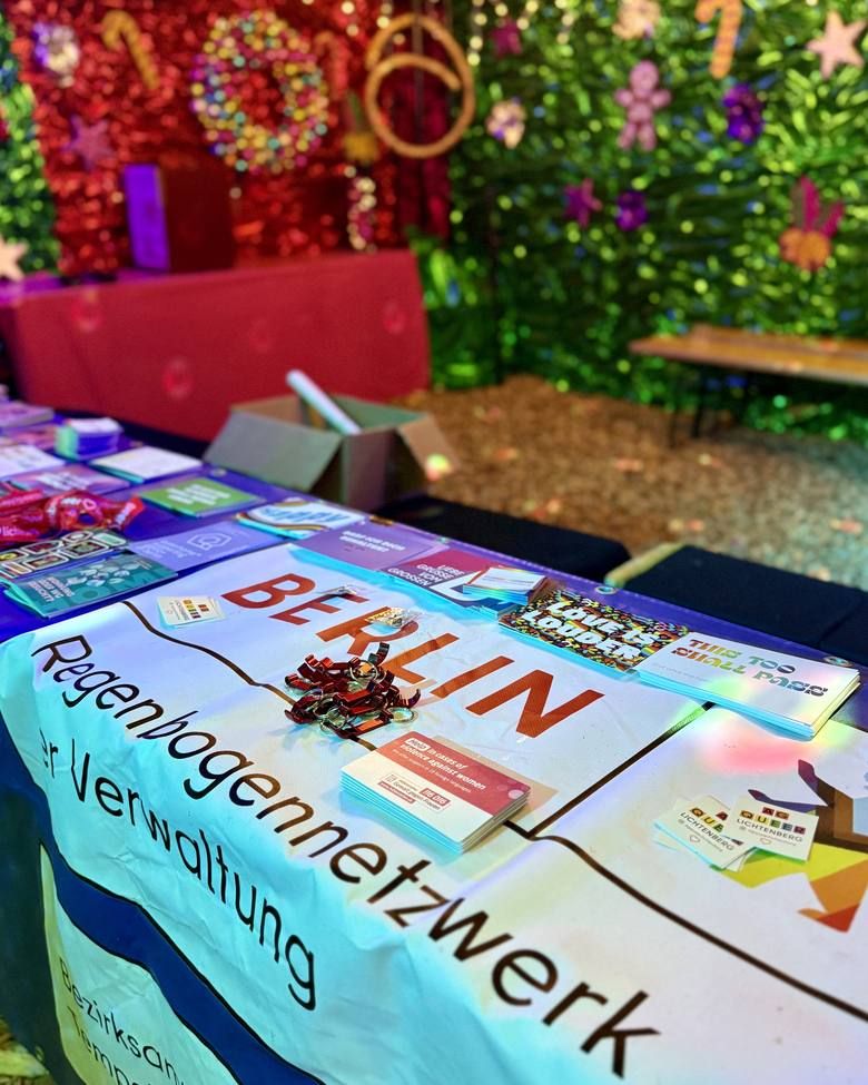 Ein Infostand des Regenbogennetzwerks Berlin präsentiert Flyer und Informationsmaterial auf einem Tisch vor einer festlich geschmückten Kulisse.
