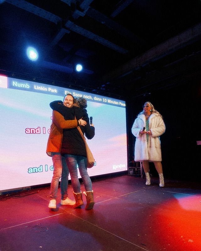 Ein Paar umarmt sich auf einer Bühne während eines Karaoke-Auftritts, während eine Dragqueen daneben auf der Bühne steht.
