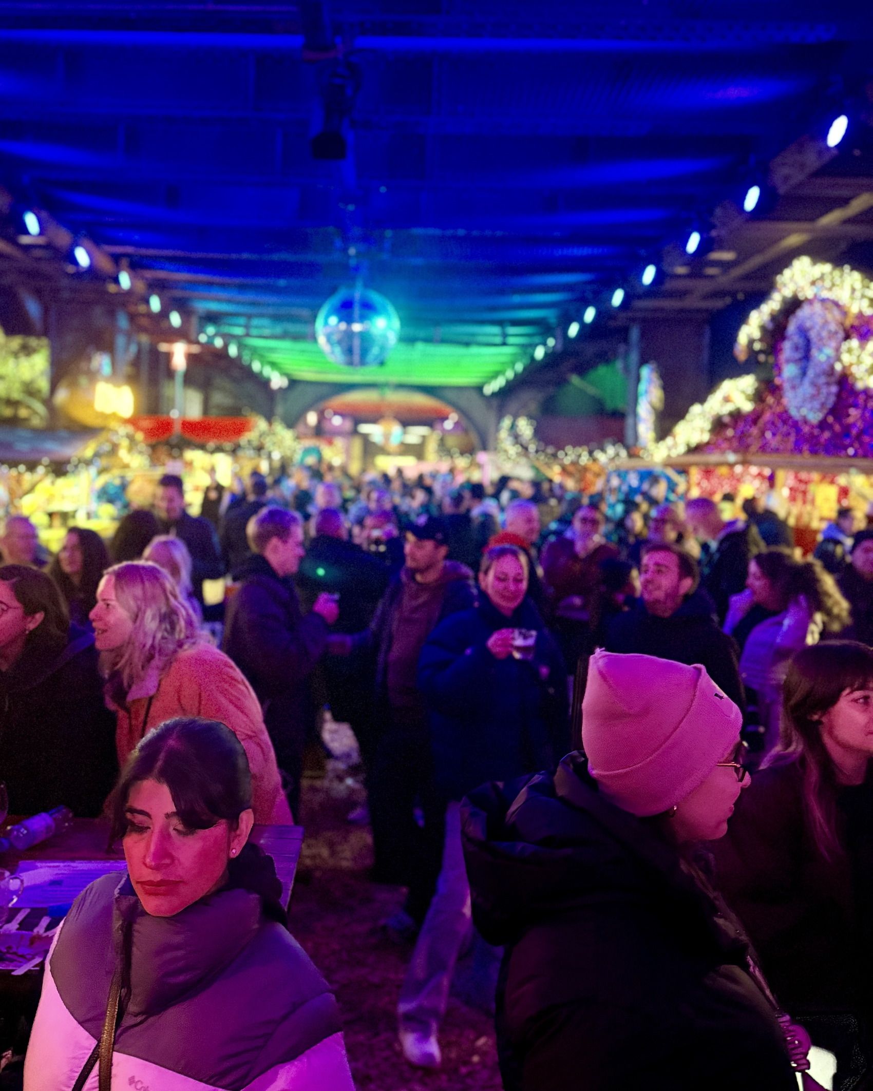 Eine dicht gedrängte Menschenmenge feiert fröhlich bei bunter Beleuchtung und Discokugel unter einer überdachten Markthalle oder einem Eventgelände.