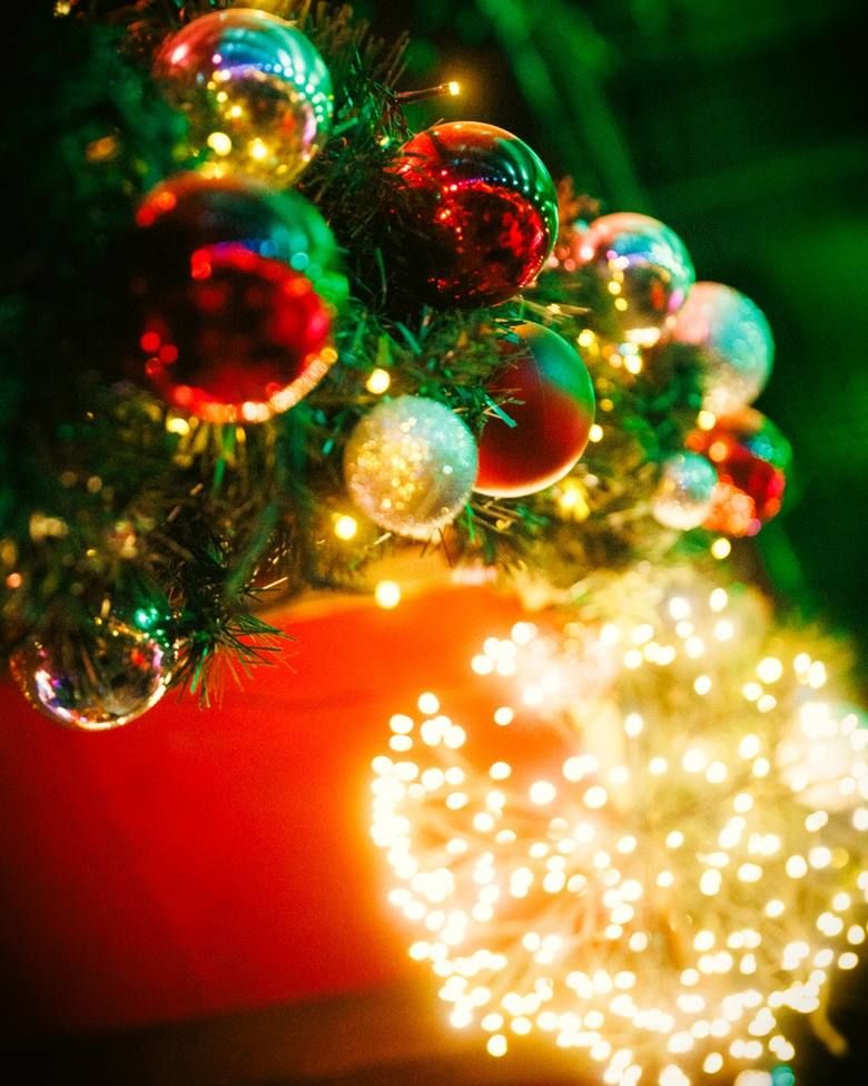 Bunte Weihnachtskugeln hängen an einem geschmückten Tannenbaum, während im Vordergrund eine helle Lichterkette für festliche Stimmung sorgt.