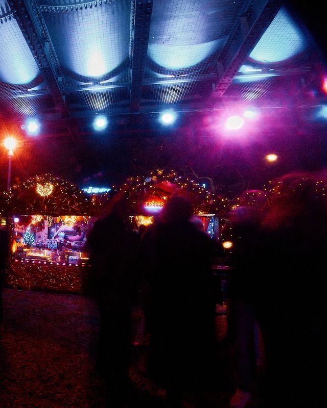 Besucher stehen bei Nacht vor einem festlich beleuchteten Crêpes-Stand auf einem stimmungsvollen Weihnachtsmarkt unter einer Brückenkonstruktion.