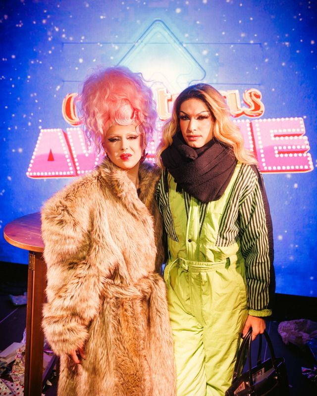 Zwei Drag-Künstlerinnen in auffälligen Outfits stehen vor einem beleuchteten Banner auf der Christmas Avenue Bühne.