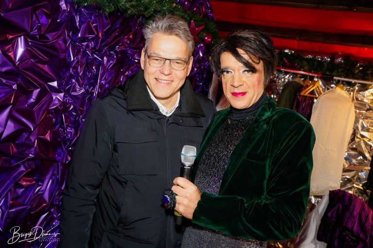 Bezirksbürgermeister von Tempelhof-Schöneberg, Jörn Oltmann, steht links im Bild und lächelt neben Marie Mondieu, die rechts im Bild ein Mikrofon hält, bei der Eröffnung der Christmas Avenue, einem LGBTQI-Weihnachtsmarkt am Nollendorfplatz in Berlin